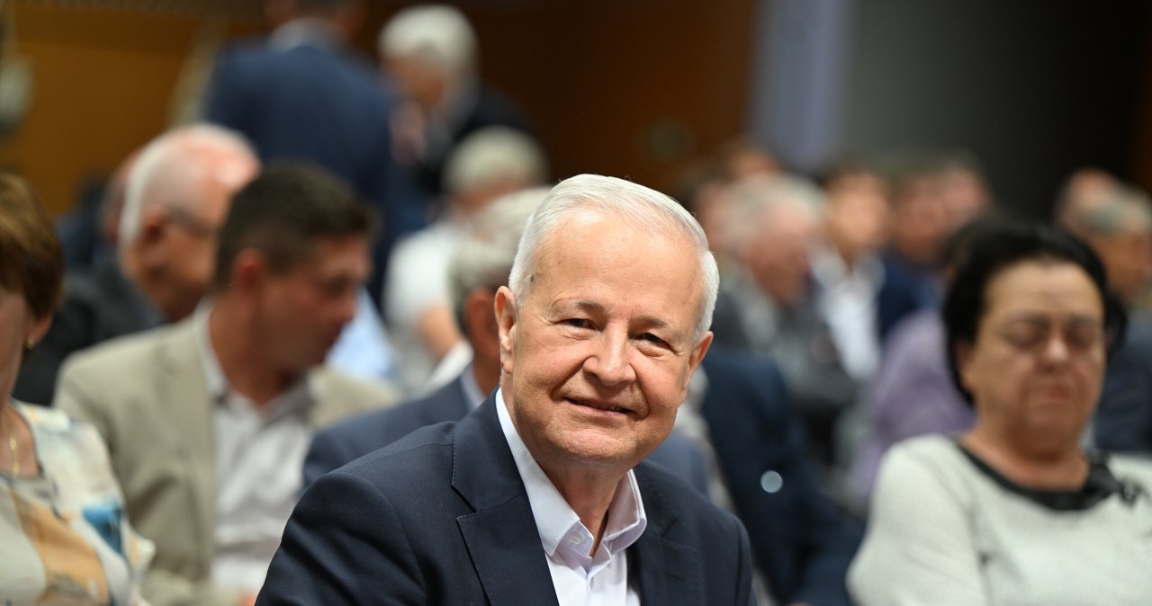 Apoloniusz Tajner, ubrany w garnitur, uśmiecha się podczas konferencji prasowej, trzymając mikrofon z logo PZN.