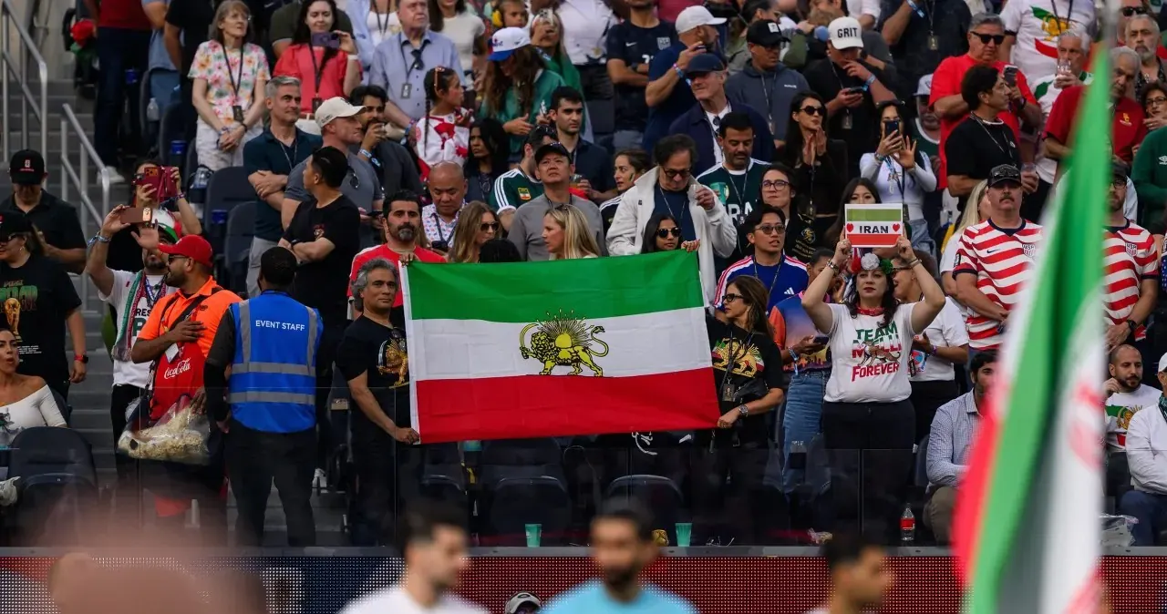 Kibice gwiżdżący podczas odgrywania hymnu Iranu na stadionie SoFi w Inglewood, z flagami protestacyjnymi widocznymi w tle.