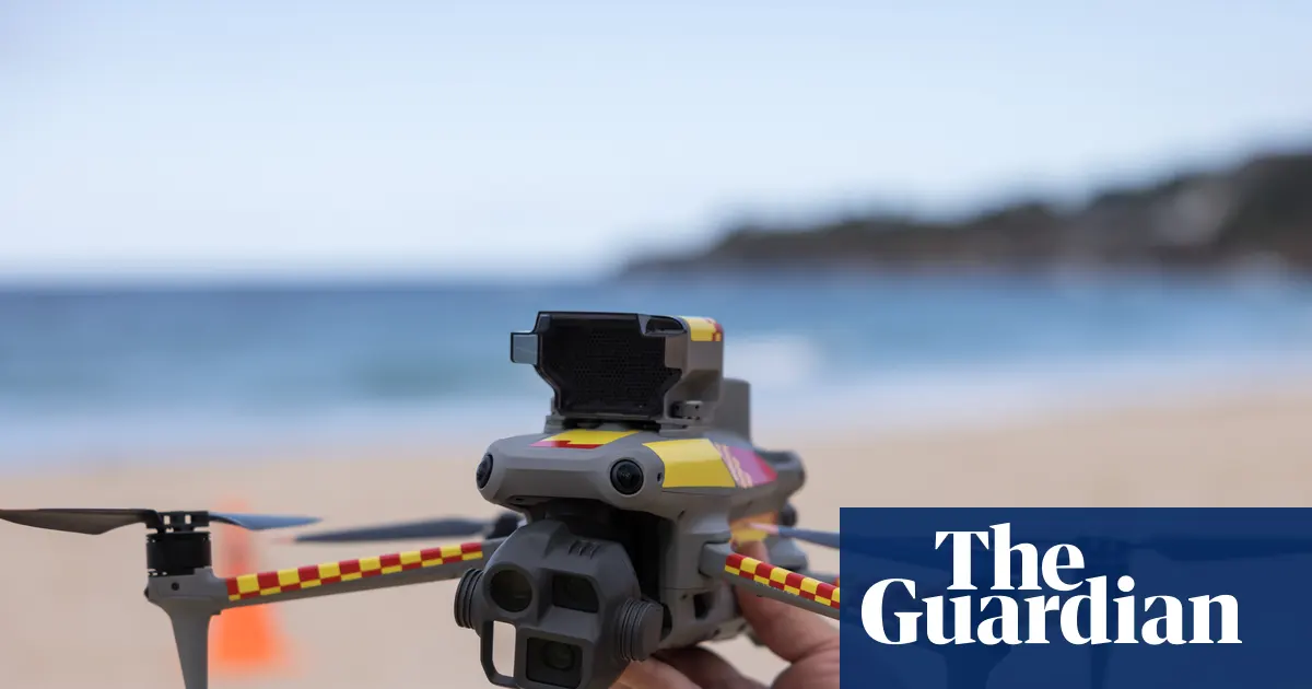 Dron latający nad australijską plażą, monitorujący wodę w poszukiwaniu rekinów, z ludźmi kąpiącymi się w oddali.