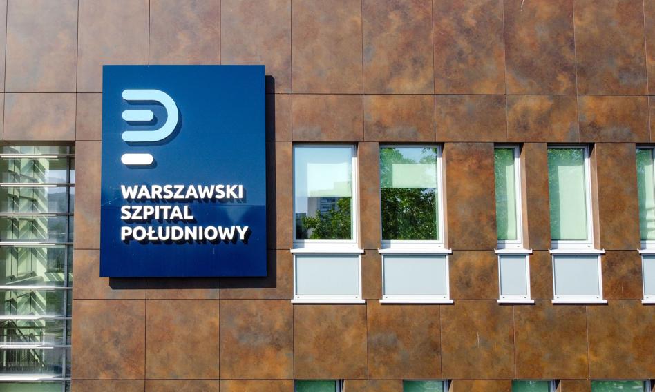 Zdjęcie przedstawiające wejście do Szpitala Południowego w Warszawie, z tabliczką informacyjną lub widocznym fragmentem budynku.
