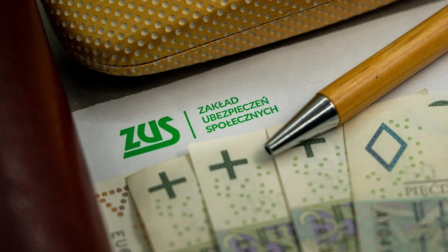 Kobieta zamyślona, przeglądająca dokumenty ZUS i bankowe, obok stos monet i kalkulator, symbolizujące nierozwiązane kwestie finansowe po zmarłej osobie.