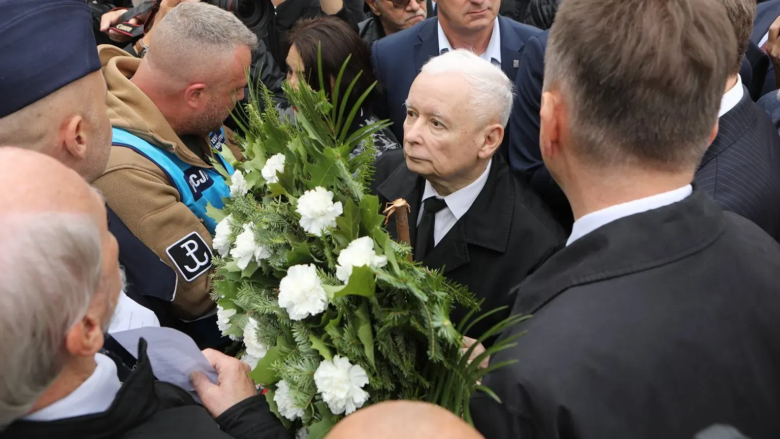 Jarosław Kaczyński, prezes Prawa i Sprawiedliwości, w otoczeniu polityków, gestykuluje podczas wystąpienia na tle pomnika, z widocznymi w oddali funkcjonariuszami policji.